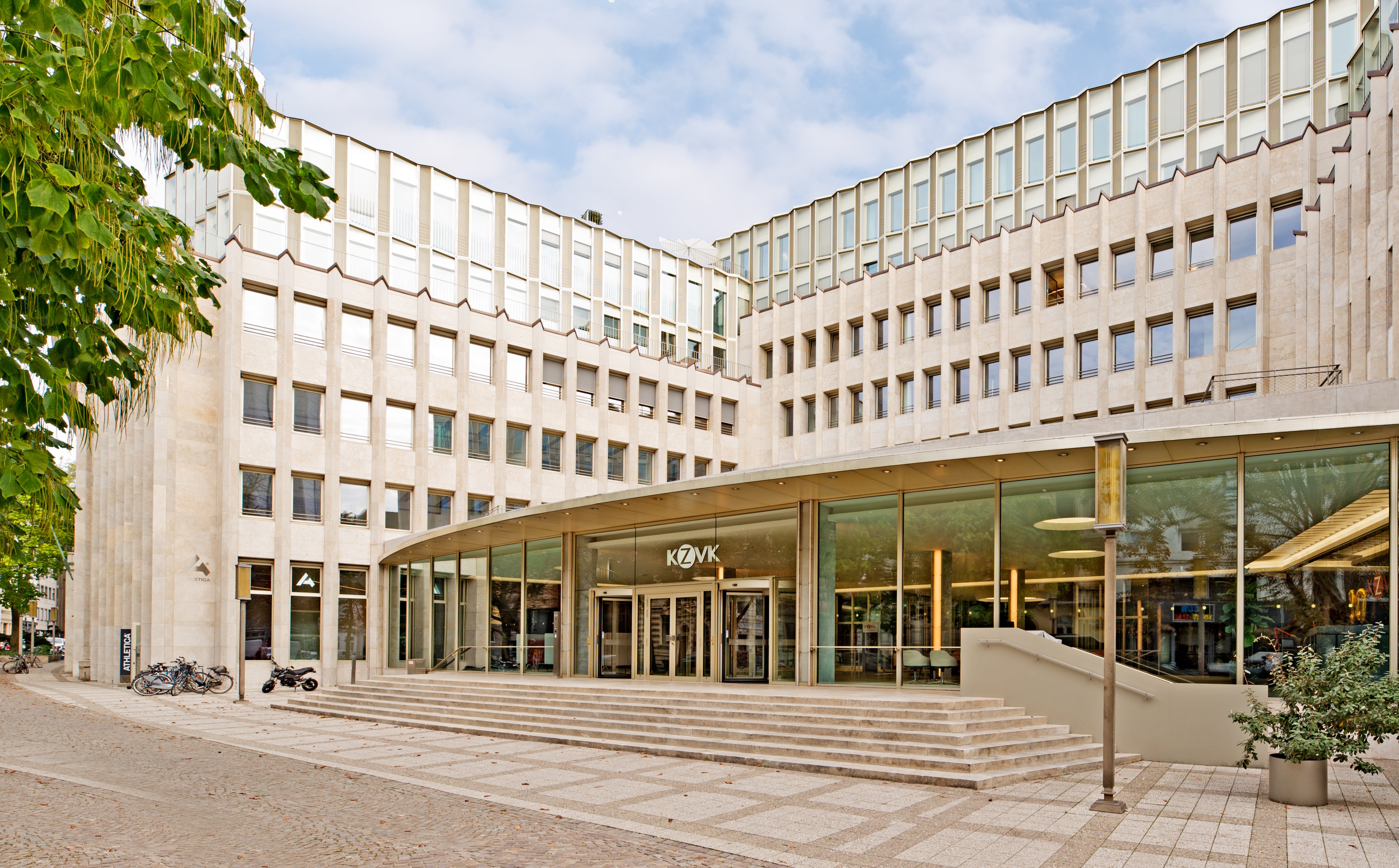 Das umgebaute Hufeisengebäude des Gerling Garden in der Kölner Altstadt-Nord mit geschwungener heller Rasterfassade, breiter Freitreppe und einem großzügigen Glas-Eingangsbereich. Die moderne Architektur integriert sich harmonisch in die historische Umgebung.