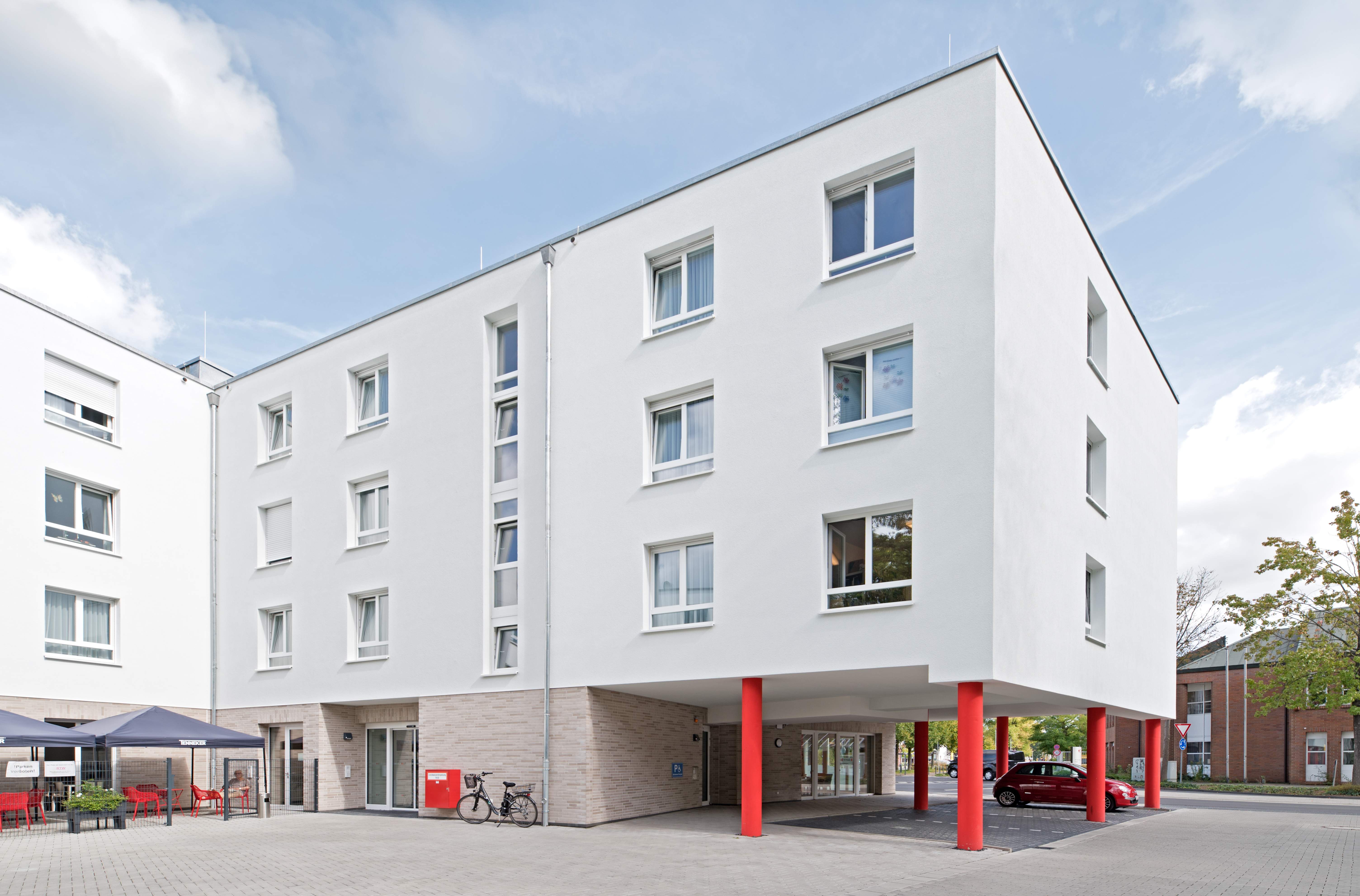 Außenansicht des dreigeschossigen Seniorenzentrums mit hellem Klinkersockel und einem überdachten Vorplatz, der von markanten roten Säulen gestützt wird.