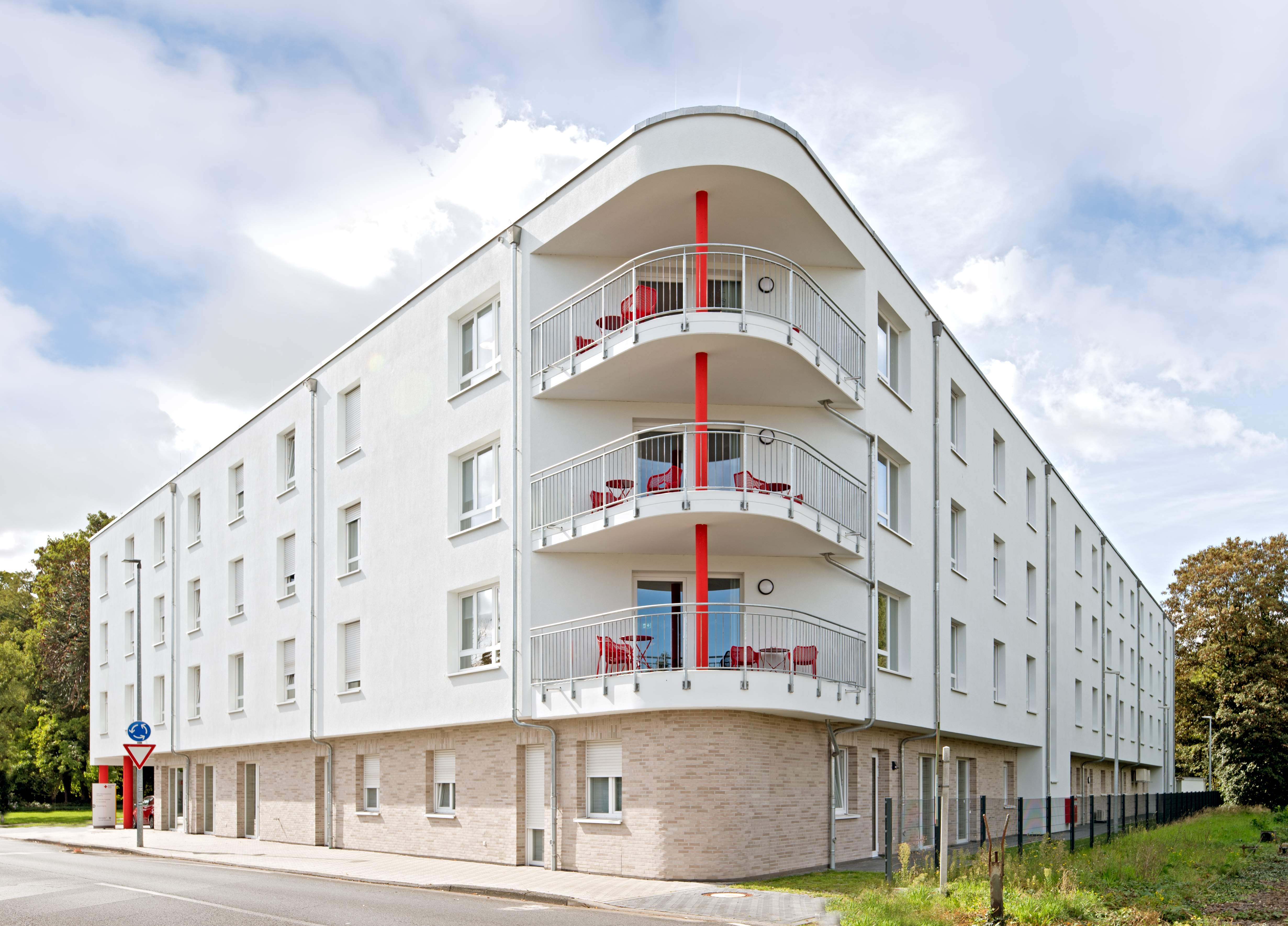 Eckansicht des Seniorenzentrums mit halbrunden Balkonen für die Bewohner der vollstationären Pflegeplätze, akzentuiert durch eine durchgehende rote Stützsäule. Die Fassade ist in einem hellen Farbton gehalten, und die Fenster sind gleichmäßig über die drei Etagen verteilt.