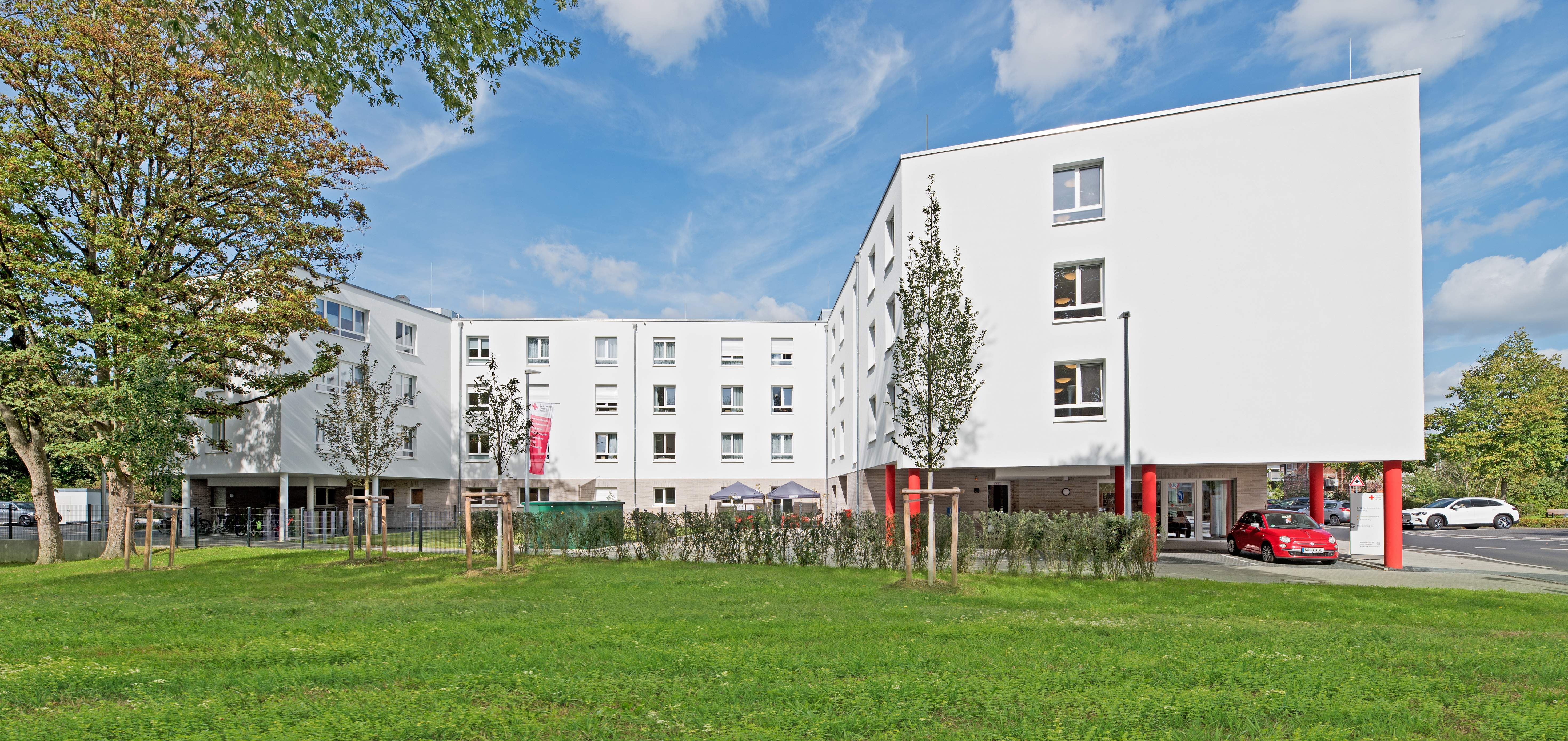 Weitwinkelaufnahme des modernen, dreigeschossigen Seniorenzentrums mit angrenzender grüner Rasenfläche und weitläufigem Außenbereich der Pflegeeinrichtung. Die Fassade ist hell, und die markanten roten Stützpfeiler heben sich deutlich ab. Im Vordergrund sind Gehwege und ein kleiner Parkplatz zu sehen, die den Zugang zum Gebäude erleichtern.