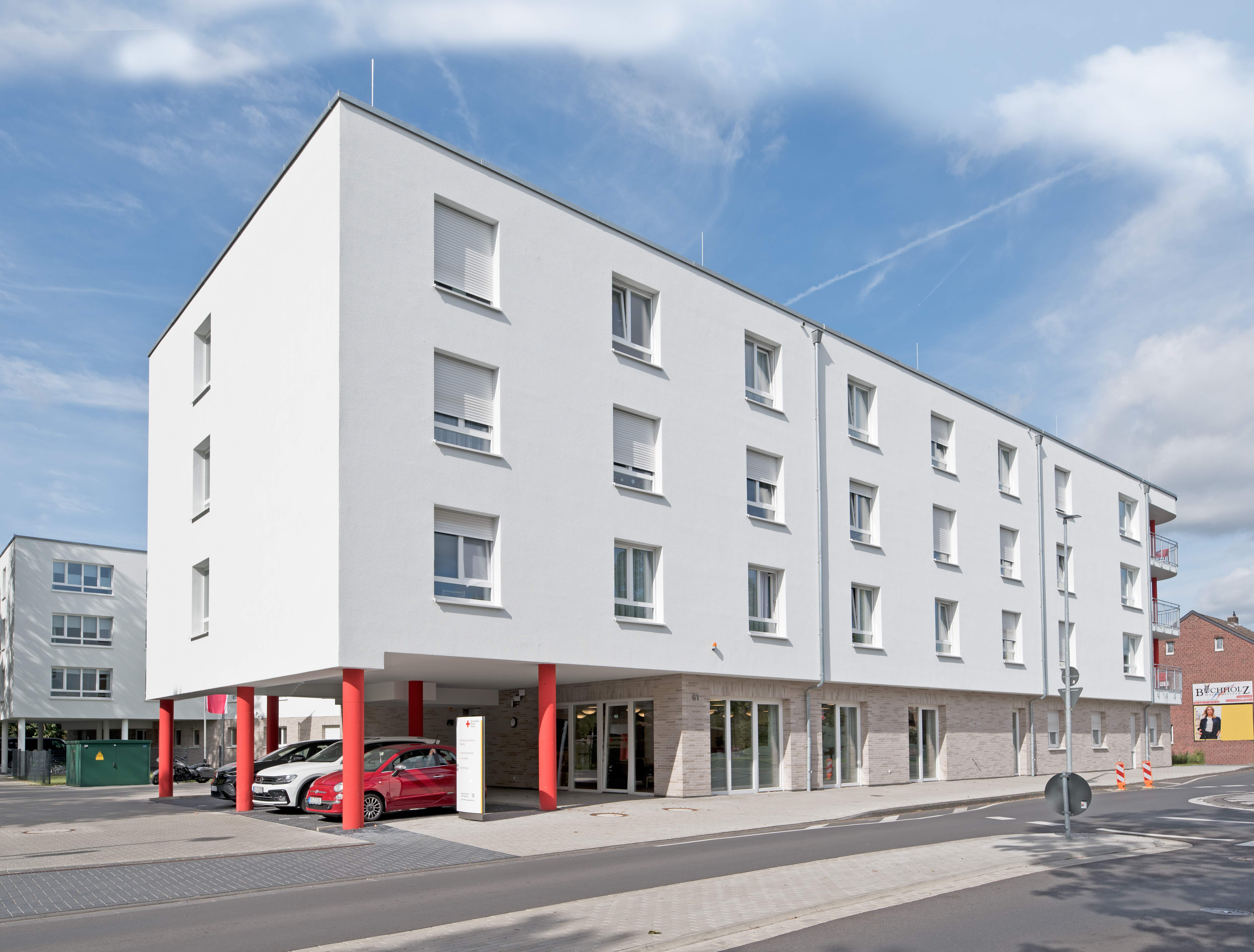 Straßen- und Eingangsansicht des dreigeschossigen Seniorenzentrums mit 84 Pflegeplätzen, dessen überhängender Trakt überdachte Parkmöglichkeiten und den Haupteingang bietet. Die Fassade ist hell, und die markanten roten Stützpfeiler heben sich deutlich ab. Im Vordergrund sind Gehwege und ein kleiner Parkplatz zu sehen, die den Zugang zum Gebäude erleichtern.