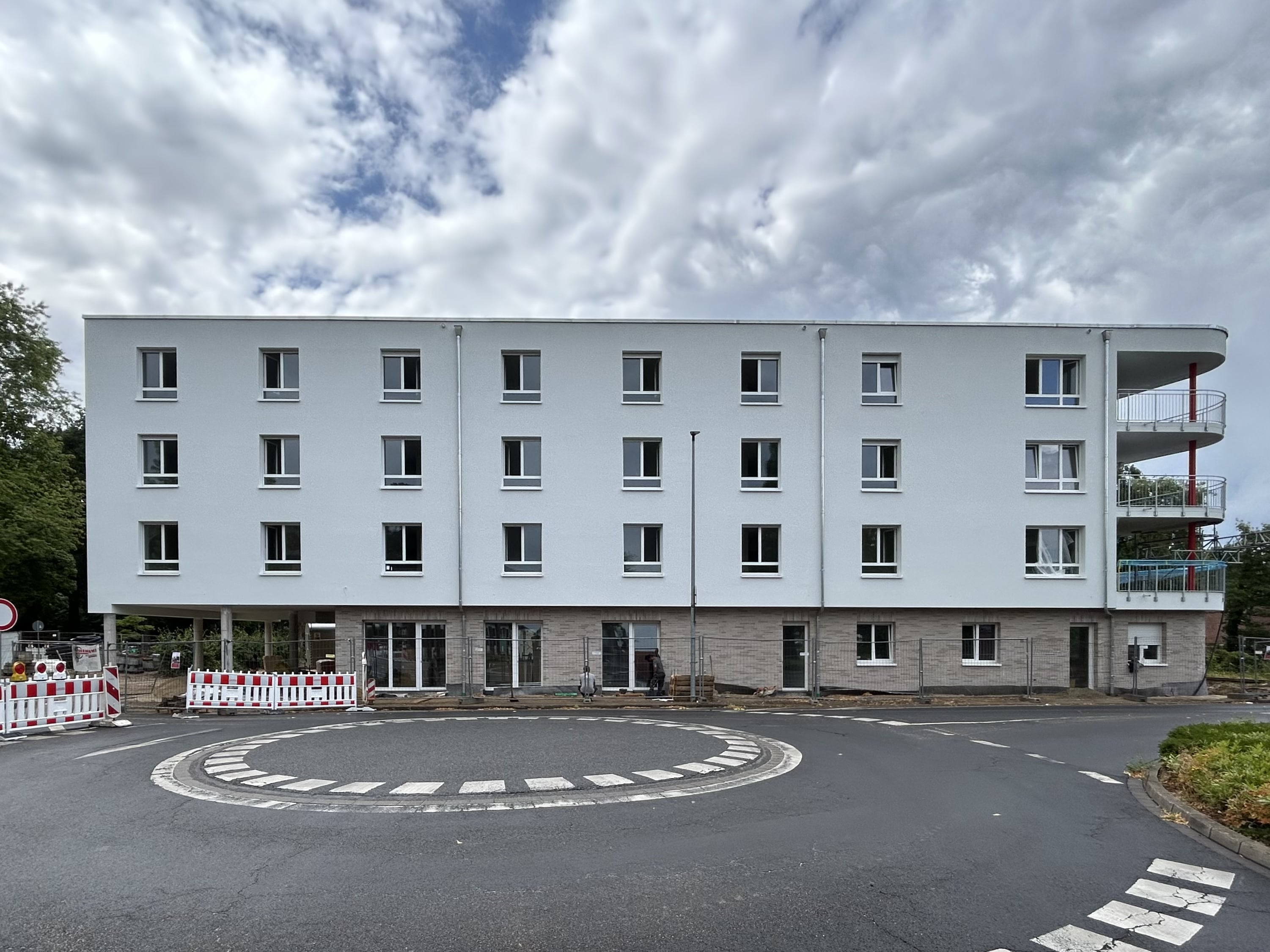 Visualisierung eines modernen Bürogebäudes an einer großen Kreuzung. Der Gebäudekomplex besteht aus einem weißen Eckgebäude mit Rundbögen und einem längeren Flügel mit vertikalen Fassadenelementen. Autos und Fußgänger auf der Straße, blauer Himmel mit Wolken.