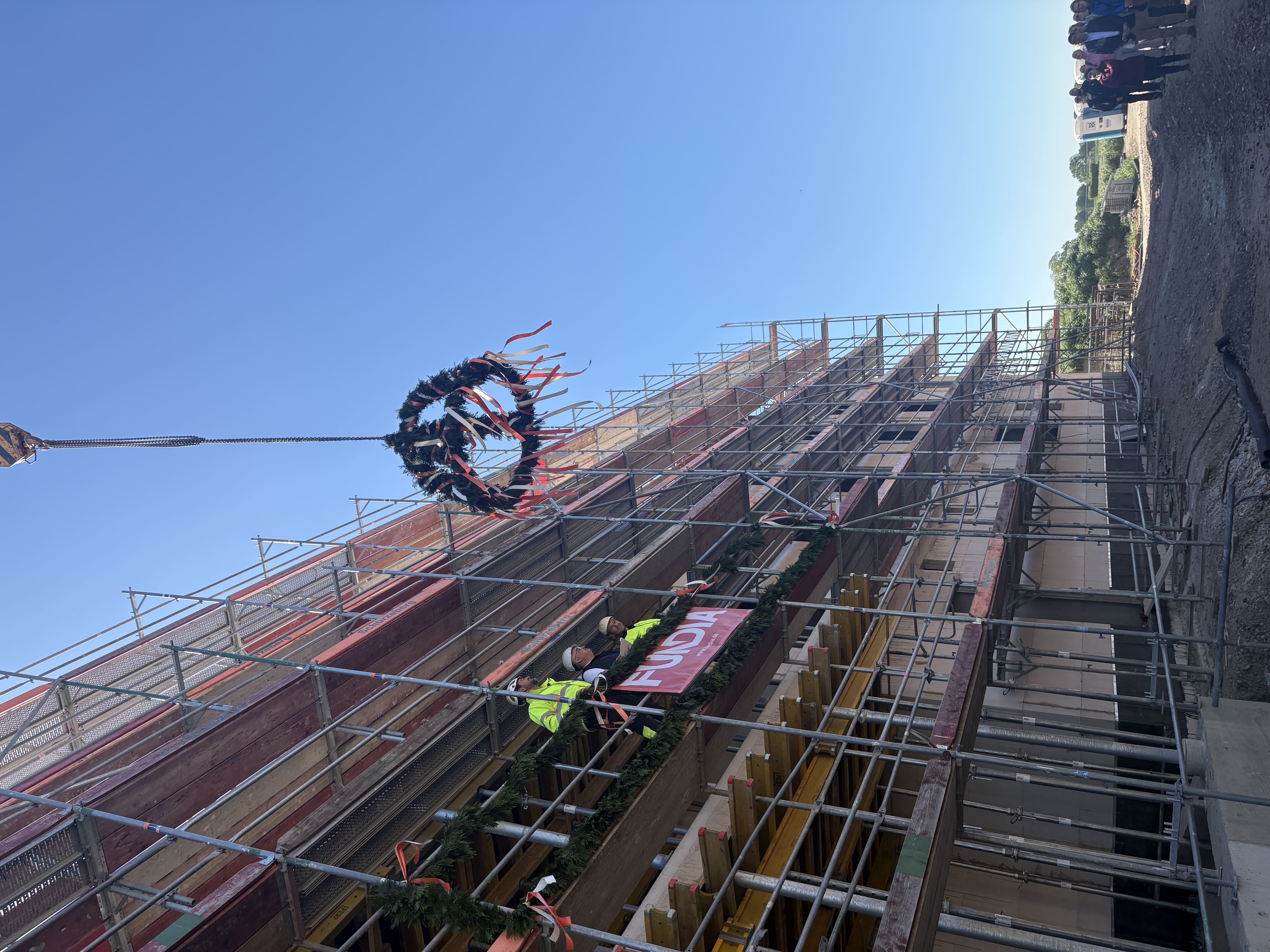 Vertikale Aufnahme der Baustelle: Ein großer, mit Bändern geschmückter Richtkranz schwebt an einem Kran über dem eingerüsteten Rohbau. Auf dem Gerüst stehen drei Bauarbeiter.