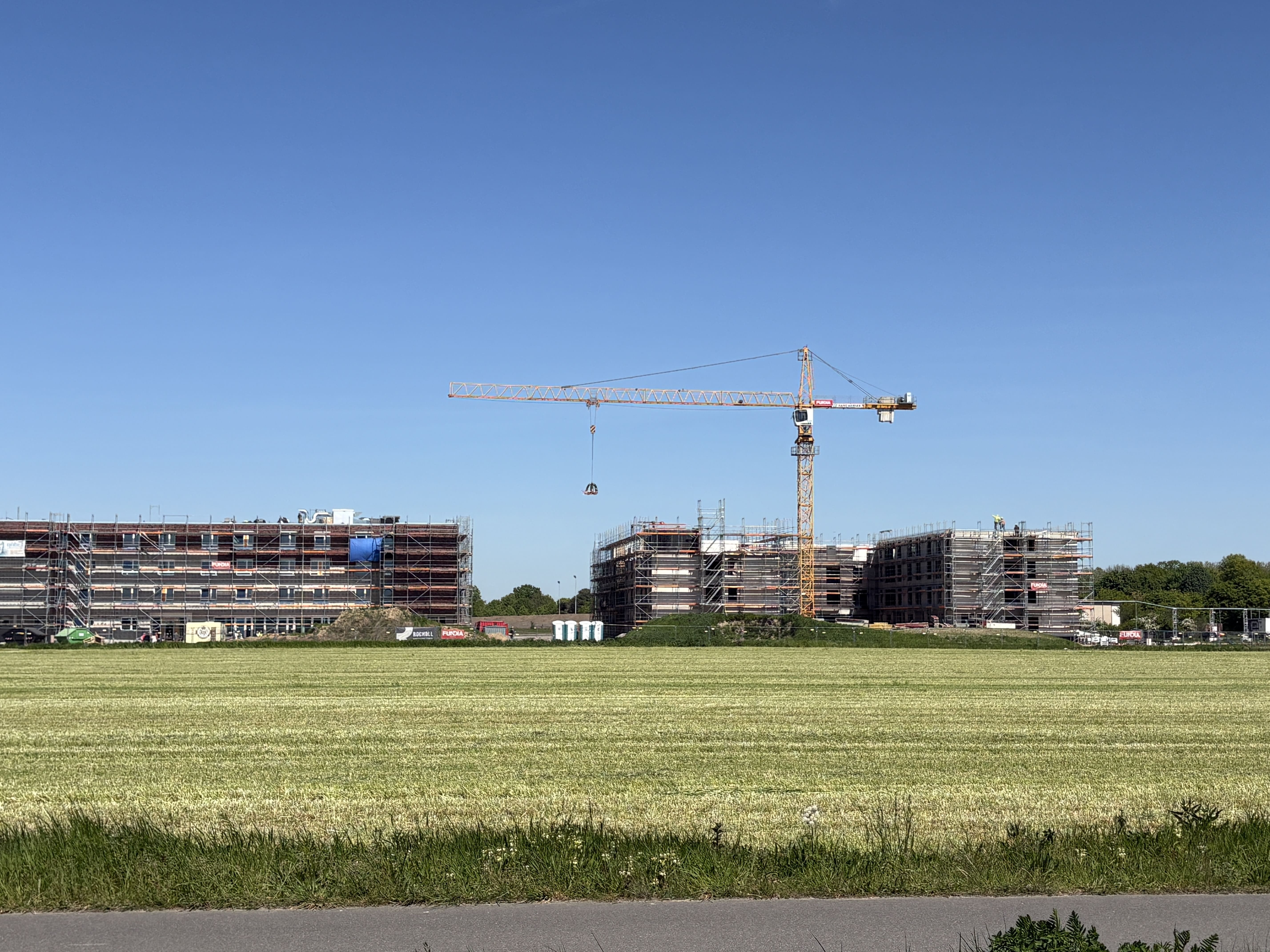 Weitwinkel-Aufnahme der Großbaustelle: Zwei ausgedehnte, eingerüstete Gebäudekomplexe in der Rohbauphase stehen hinter einer weiten, grünen Wiese. Mittig ragt ein gelber Baukran in den blauen Himmel.