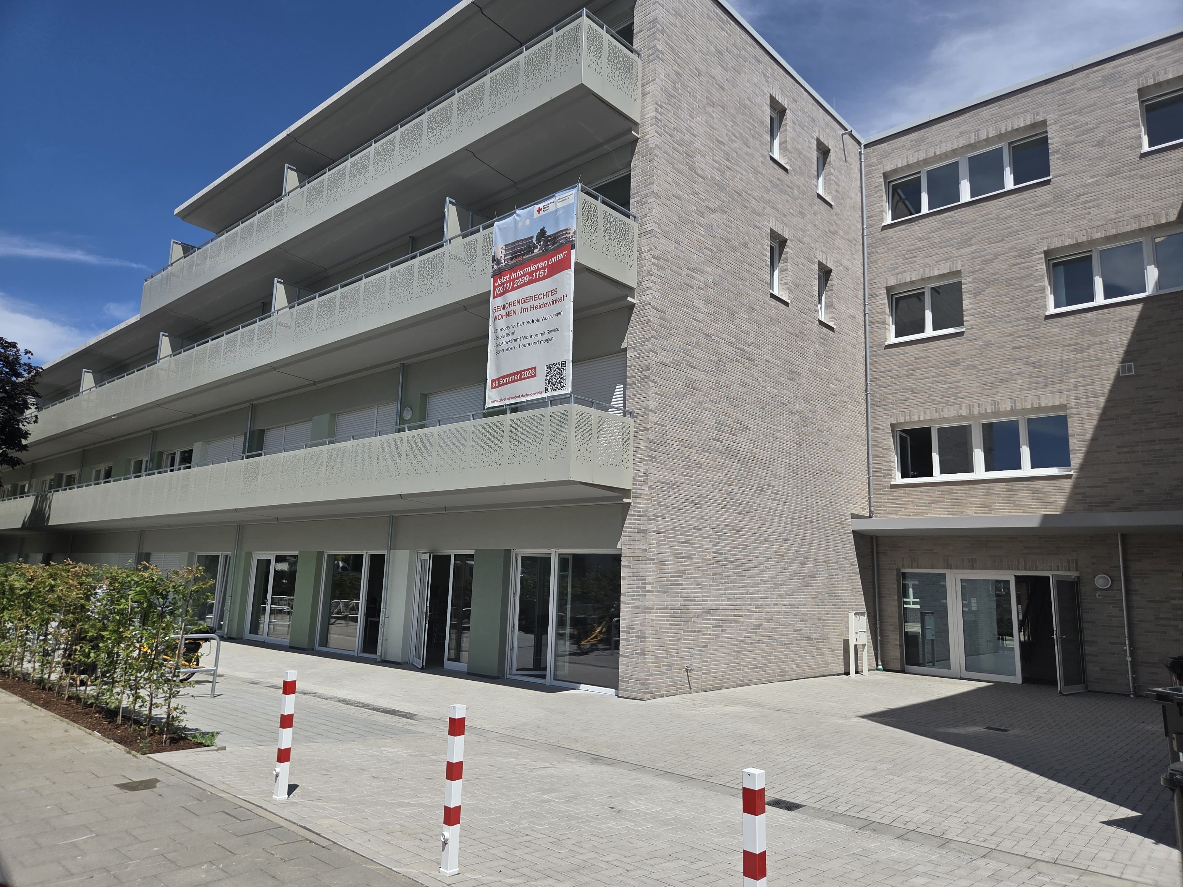 Außenansicht eines modernen, mehrstöckigen Wohngebäudes mit heller Klinkerfassade. Über mehrere Etagen der linken Balkonreihe hängt ein großes Werbebanner des Deutschen Roten Kreuzes mit der Aufschrift „SENIORENGERECHTES WOHNEN ‚Im Heiden Wolf‘“. Im Erdgeschoss befinden sich großzügige Glasfronten und offene Türen. Im gepflasterten Vordergrund ragen drei rot-weiße Absperrpfosten aus dem Boden.